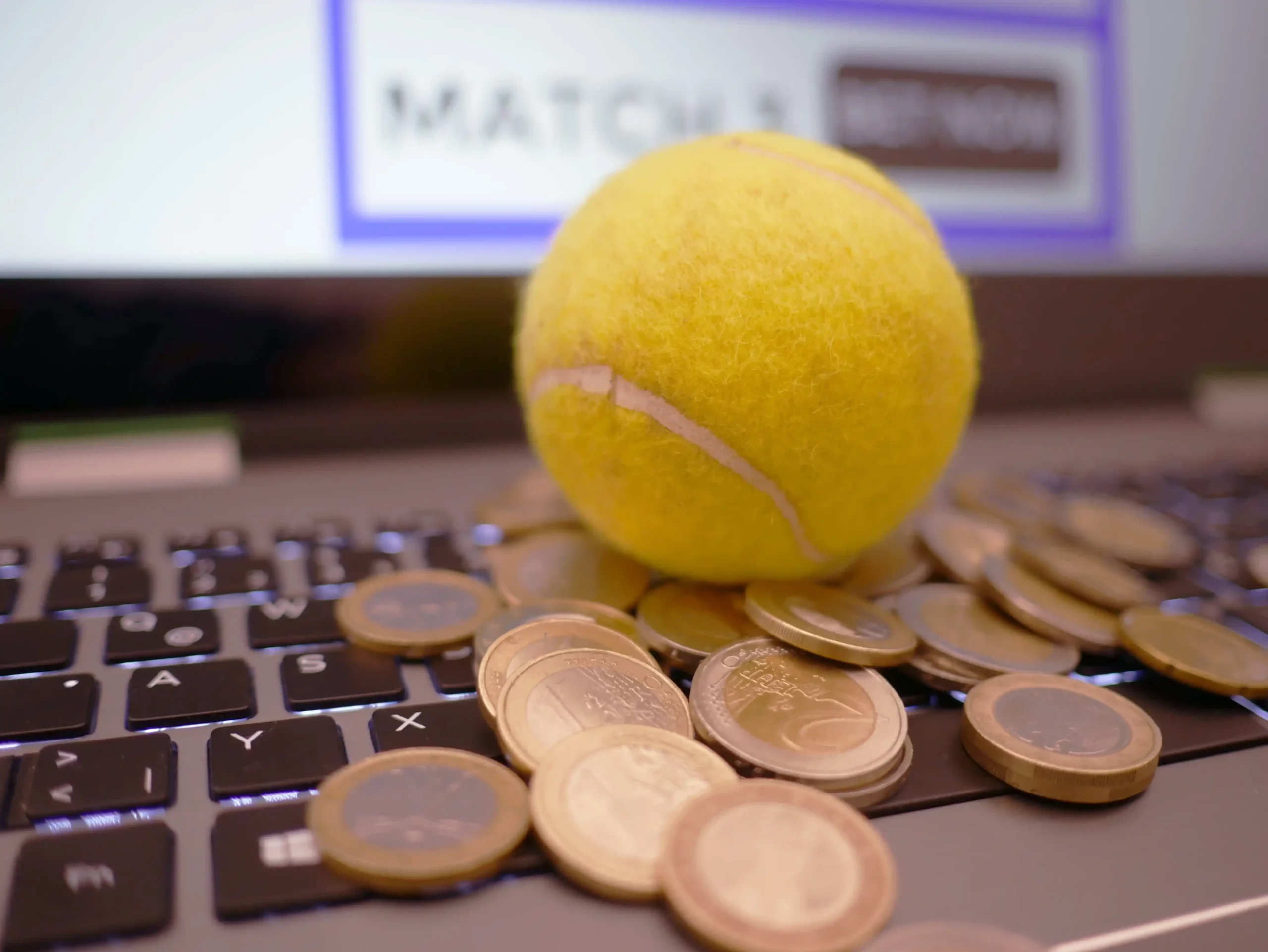 Balle de tennis, pièces de monnaie et ordinateur illustrant le lien entre sport, argent et paris sportifs en ligne.